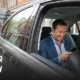 Passenger using phone in minicab while driver focuses on road during city travel